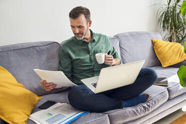 Man working from home looking at documents sitting cross-legged with laptop on sofa - BSZF02242