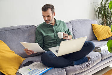 Mann, der von zu Hause aus arbeitet und im Schneidersitz mit Laptop auf dem Sofa sitzend Dokumente betrachtet - BSZF02242