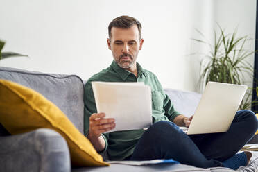 Worried man working from home looking at documents sitting with laptop on sofa - BSZF02243