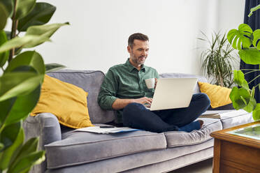 Relaxed man using laptop on the sofa while working from home - BSZF02244
