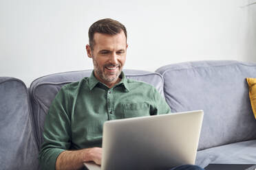 Relaxed man working from home using laptop sitting on the sofa - BSZF02246