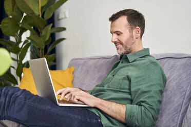 Relaxed man working from home using laptop sitting on the sofa - BSZF02247