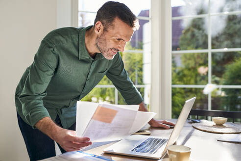 Lächelnder Mann erledigt Papierkram zu Hause am Laptop - BSZF02251
