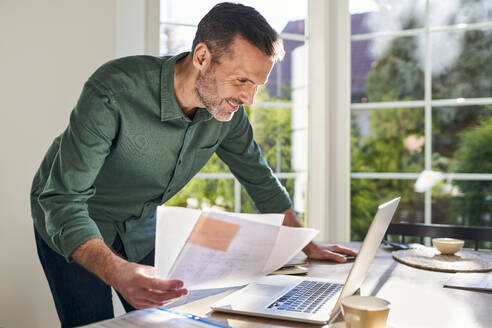 Smiling man doing paperwork at home using laptop - BSZF02251