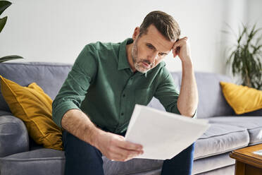 Worried man looking at documents sitting on sofa - BSZF02256
