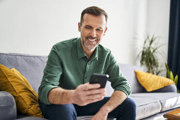 Fröhlicher Mann sitzt auf der Couch und benutzt sein Smartphone - BSZF02259