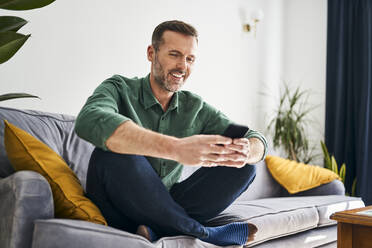 Fröhlicher Mann sitzt auf der Couch und benutzt sein Smartphone - BSZF02260