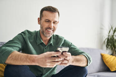 Cheerful man sitting on couch using smartphone - BSZF02261