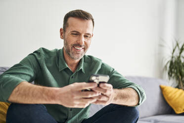 Fröhlicher Mann sitzt auf der Couch und benutzt sein Smartphone - BSZF02261