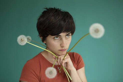 Thoughtful woman with hand on chin holding dandelions against green background - AXHF00258