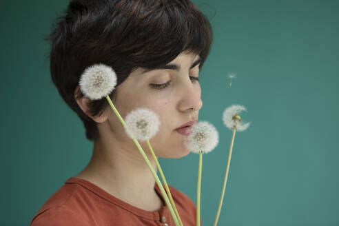 Young woman smelling dandelions against green background - AXHF00262