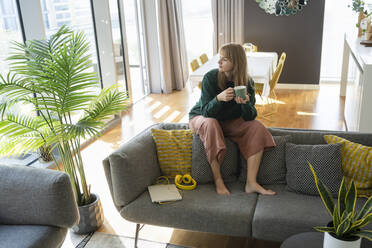 Nachdenkliche Frau mit Teetasse, die zu Hause auf dem Sofa sitzt - SVKF01236