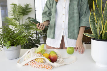Woman using smart phone and bag of fresh fruits and vegetables at home - SVKF01250
