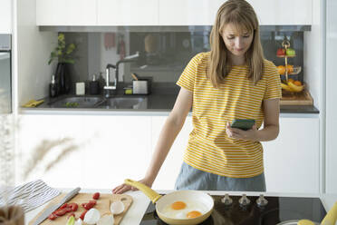 Frau macht Spiegeleier und benutzt Smartphone zu Hause - SVKF01263