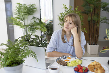 Contemplative freelancer sitting with breakfast and laptop at desk - SVKF01266