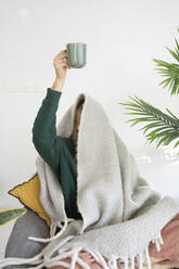 Woman holding coffee cup sitting under blanket at home - SVKF01277