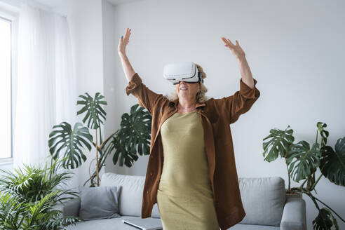 Sorglose Frau mit erhobenen Armen, die ein Virtual-Reality-Headset trägt und zu Hause steht - AAZF00008