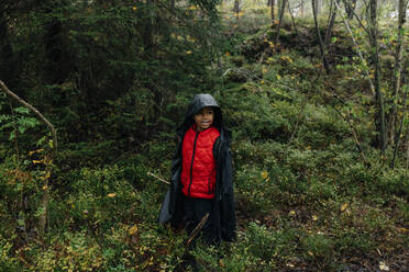 Smiling boy in raincoat looking away while standing amidst plants in forest - MASF34672