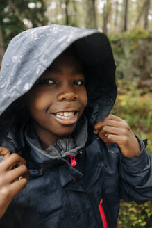 Portrait of boy with toothy smile holding hood of raincoat in forest - MASF34673
