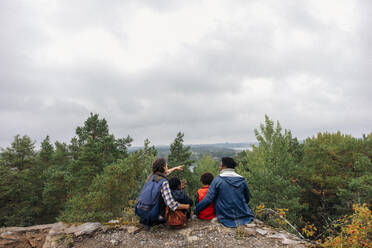 Rückansicht einer Frau, die ihrer Familie Bäume zeigt, während sie auf einer Klippe gegen den Himmel sitzt - MASF34694