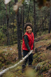 Smiling elementary boy picking up firewood while standing in forest - MASF34695
