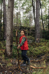 Seitenansicht eines Jungen, der wegschaut und Brennholz bei Bäumen im Wald hält - MASF34704