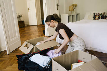 Seitenansicht einer Frau, die zu Hause kniend Kleidung in einen Karton packt - MASF34733