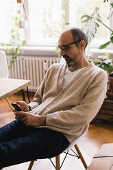 Mann benutzt Smartphone, während er zu Hause auf einem Stuhl sitzt - MASF34767