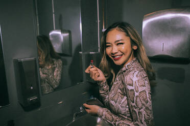 Portrait of smiling young woman applying lipstick in bathroom at nightclub - MASF34832