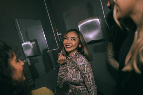 Lächelnde junge Frau, die Lippenstift aufträgt, während sie ihre Freunde im Badezimmer eines Nachtclubs ansieht - MASF34833