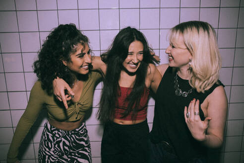Fröhliche junge, gemischtrassige Freundinnen, die mit den Armen an einer weißen Wand in einem Nachtclub stehen - MASF34842