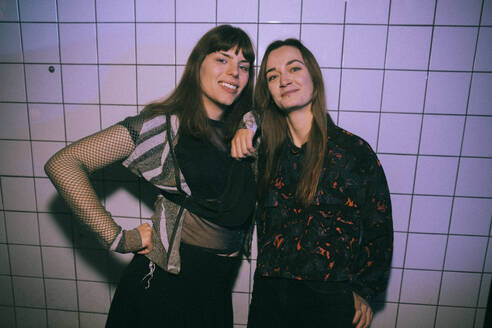 Porträt einer lächelnden Transgender-Frau mit einer Freundin, die in einem Nachtclub an einer weißen Wand steht - MASF34843