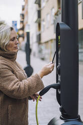 Ältere Frau, die an einer Ladestation für Elektrofahrzeuge mit einer digitalen Geldbörse bezahlt - MASF34914