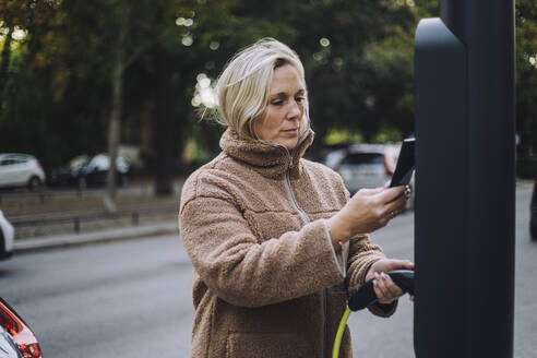 Frau hält ein Ladegerät in der Hand, während sie ihr Smartphone an einer Ladestation durchsucht - MASF34915