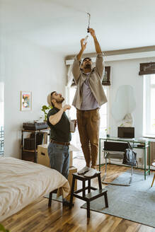 Mann installiert Glühbirne auf einem Hocker stehend bei seinem Freund zu Hause - MASF34927