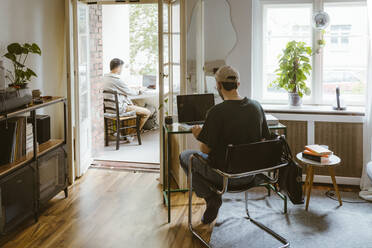 Rückansicht eines schwulen Paares, das zu Hause auf einem Stuhl sitzend Laptops benutzt - MASF34939