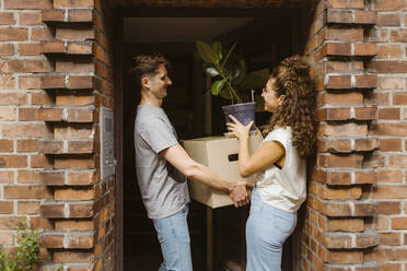 Glückliches Paar mit Pappkarton und Topfpflanze, das am Eingang miteinander spricht - MASF34956