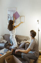 Side view of man looking at girlfriend mounting photo frame on wall - MASF34959