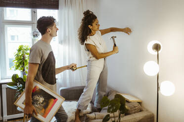 Woman hammering nail in wall with boyfriend holding picture frame and measuring tape at home - MASF34963