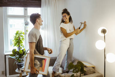 Happy girlfriend with hammer looking at boyfriend holding frame at home - MASF34964