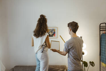 Rear view of boyfriend and girlfriend helping each other while mounting frame on wall at home - MASF34965