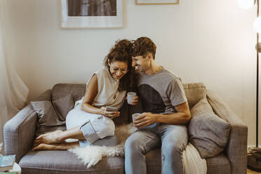 Happy couple having coffee while sharing smart phone sitting on sofa at home - MASF34972