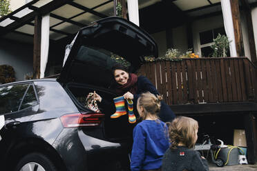 Glückliche Mutter mit Töchtern, die Stiefel und Tasche in den Kofferraum eines Elektroautos laden - MASF34981
