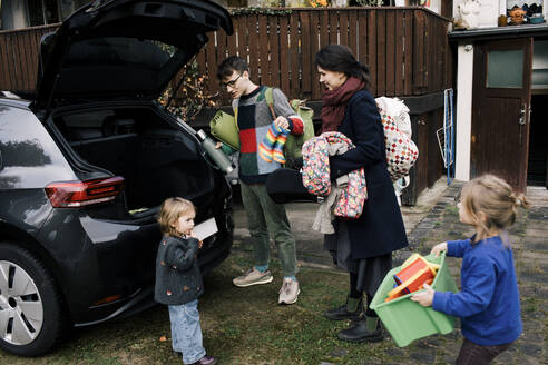 Eltern mit Kindern beim Einladen von Gepäck in den Kofferraum eines Elektroautos im Hinterhof - MASF34982