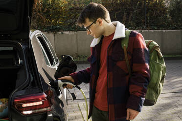 Man wearing backpack while charging electric car at station - MASF35003