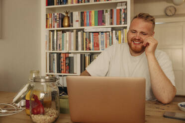 Smiling man watching laptop while sitting at home - MASF35021