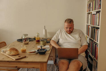 Man using laptop while sitting with breakfast by bookshelf at home - MASF35025