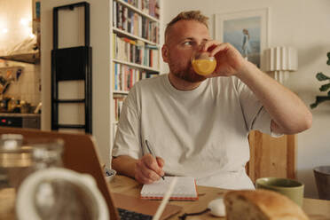 Contemplative man drinking juice while writing in diary at home - MASF35027