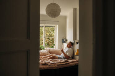 Obese man reading book in bedroom seen through doorway - MASF35034