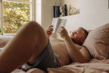 Hemdloser übergewichtiger Mann liest ein Buch, während er zu Hause auf dem Bett liegt - MASF35035